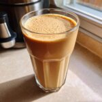 A tall glass filled with a creamy, frothy Banana Peanut Butter Smoothie sitting on a sunlit countertop.