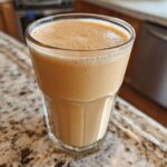 A glass filled with a creamy, light brown Banana Peanut Butter Smoothie resting on a granite countertop.