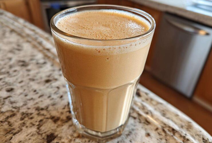 A glass filled with a creamy, light brown Banana Peanut Butter Smoothie resting on a granite countertop.