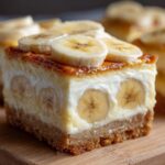 A close-up of a thick slice of Banana Pudding Cheesecake Bars showing the graham cracker crust, creamy filling with banana slices inside, and topped with fresh banana slices.