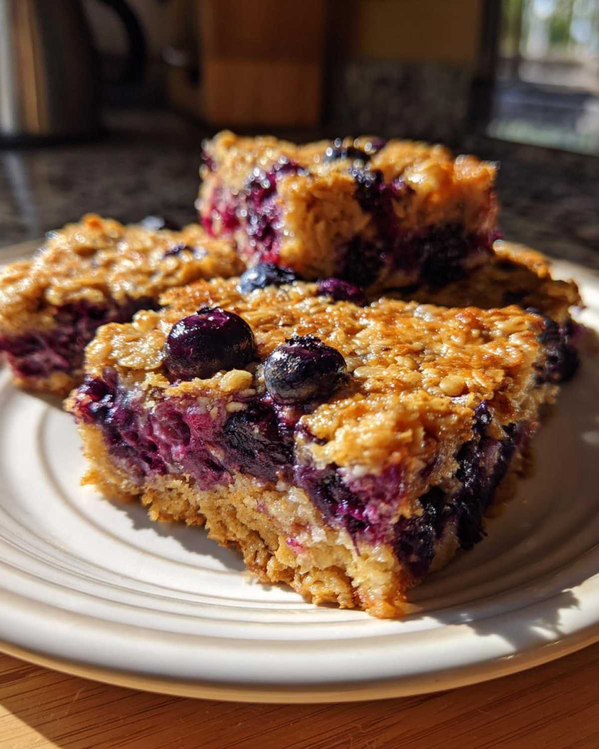 Close-up of three golden Blueberry Baked Oatmeal Squares stacked on a white plate, bursting with purple blueberries.