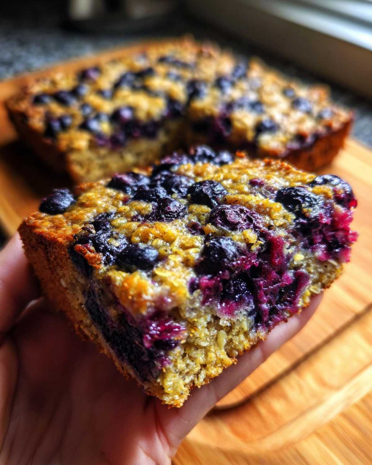 A hand holds a thick slice of moist Blueberry Baked Oatmeal Squares bursting with purple fruit.
