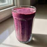 A glass filled with a thick, vibrant purple Blueberry Lemon Chia Smoothie, sitting on a counter near a window.