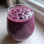 A close-up of a thick, purple Blueberry Lemon Chia Smoothie in a clear glass, topped with whole blueberries and chia seeds.