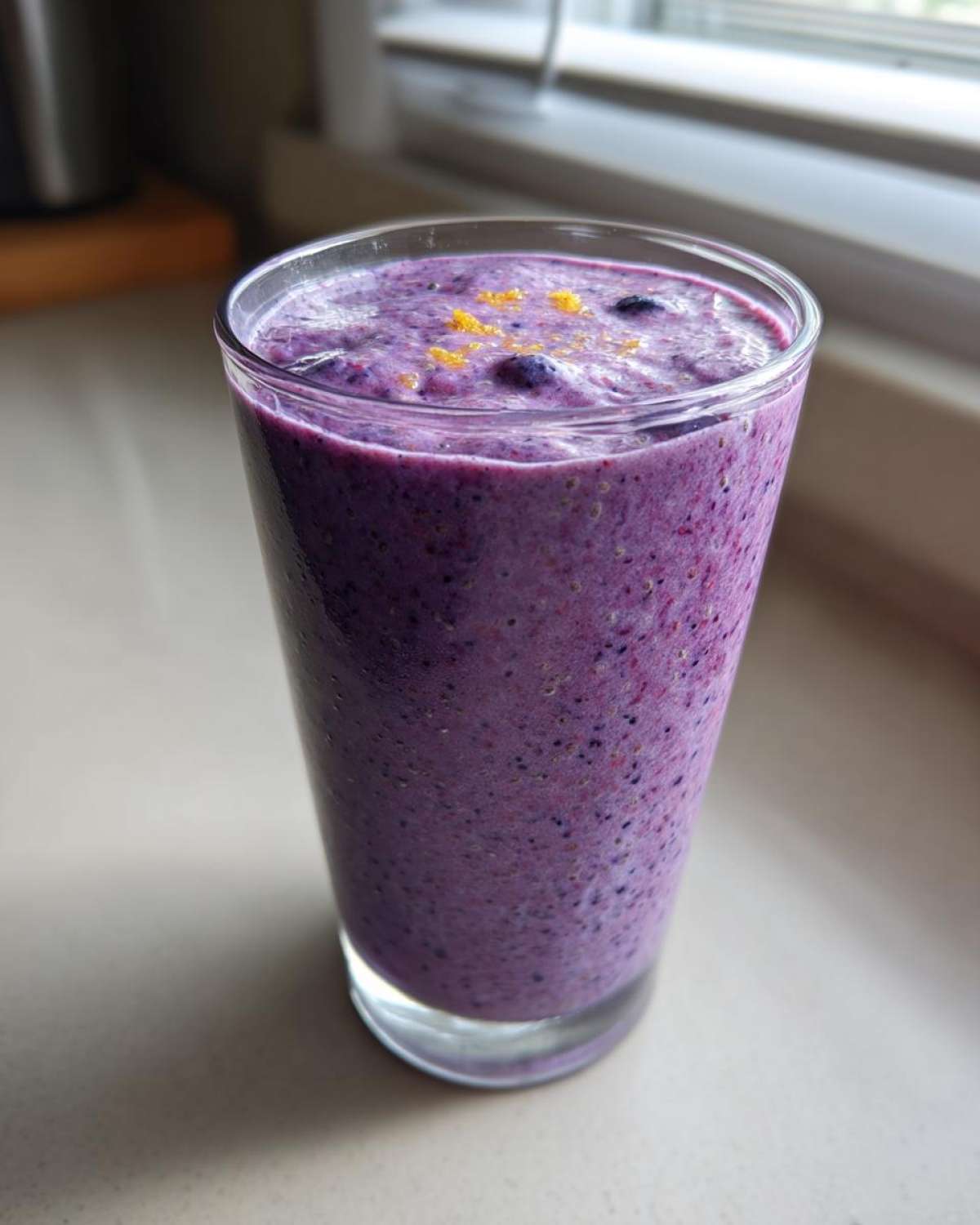 A tall glass filled with a thick, vibrant purple Blueberry Lemon Chia Smoothie, topped with zest.
