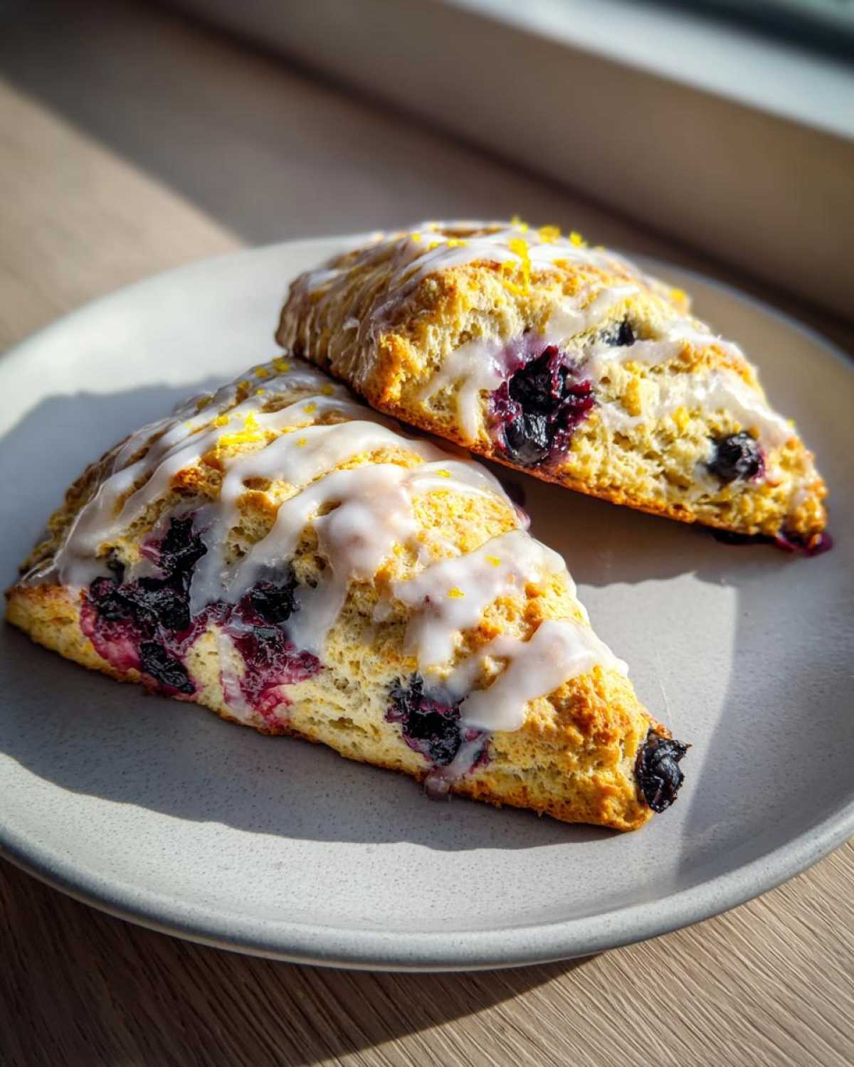 Two freshly baked Blueberry Lemon Scones topped with white glaze and lemon zest, sitting on a gray plate.