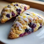 Two triangular Blueberry Lemon Scones drizzled with white glaze and topped with lemon zest on a white plate.