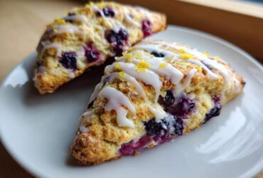 Two triangular Blueberry Lemon Scones drizzled with white glaze and topped with lemon zest on a white plate.