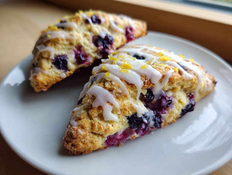 Two triangular Blueberry Lemon Scones drizzled with white glaze and topped with lemon zest on a white plate.