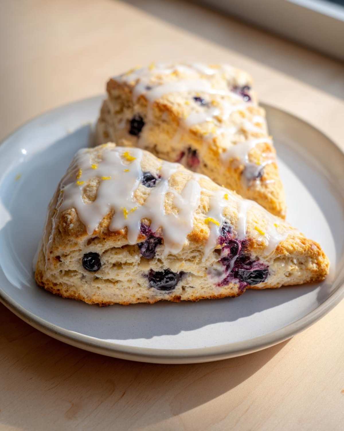 Two freshly baked Blueberry Lemon Scones topped with white glaze and lemon zest, resting on a light gray plate.