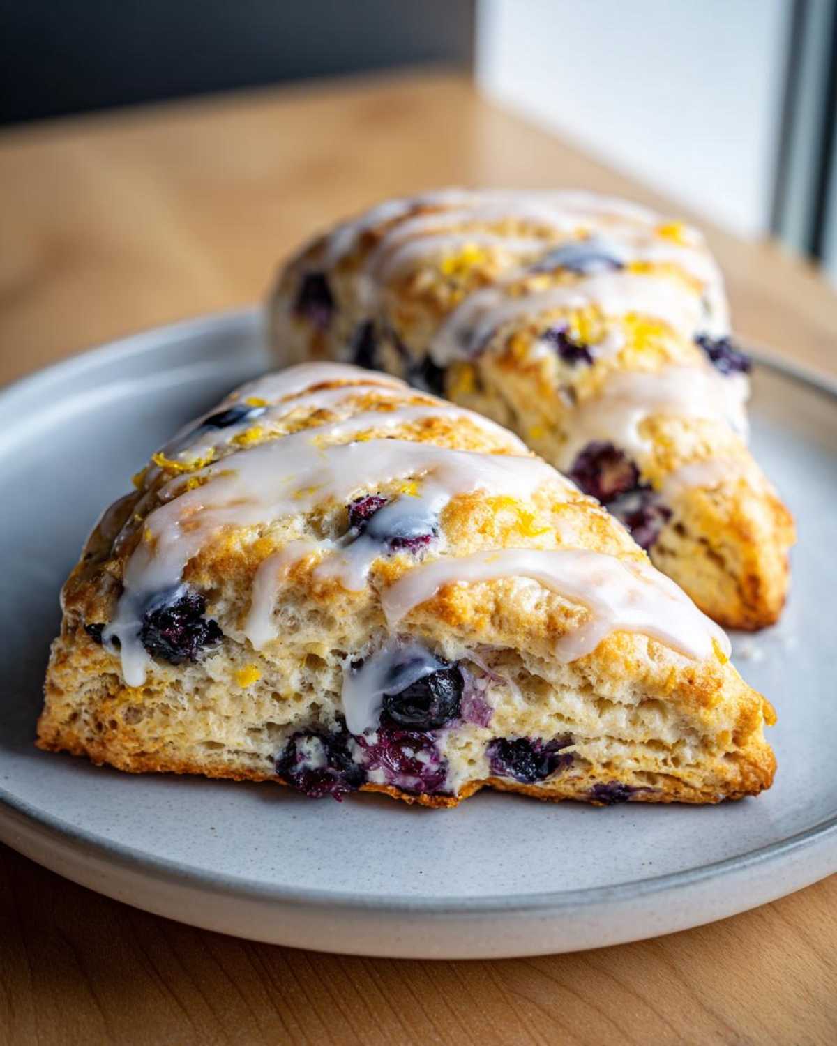 Two freshly baked Blueberry Lemon Scones topped with a sweet white glaze, sitting on a light gray plate.