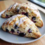 Three freshly baked Blueberry Lemon Scones drizzled with white icing and lemon zest on a gray plate.