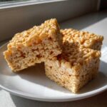Three gooey, golden Brown Butter Rice Krispie Treat Bars stacked slightly on a white plate near a window.