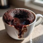 A close-up of a freshly baked Chocolate Lava Mug Cake overflowing with melted chocolate sauce in a white mug.