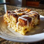 A close-up of a serving of Cinnamon Roll Overnight French Toast Bake drizzled with white icing on a white plate.