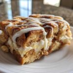 A close-up of a single serving of Cinnamon Roll Overnight French Toast Bake drizzled with white icing on a white plate.