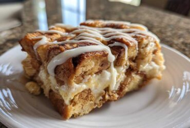 A close-up of a single serving of Cinnamon Roll Overnight French Toast Bake drizzled with white icing on a white plate.