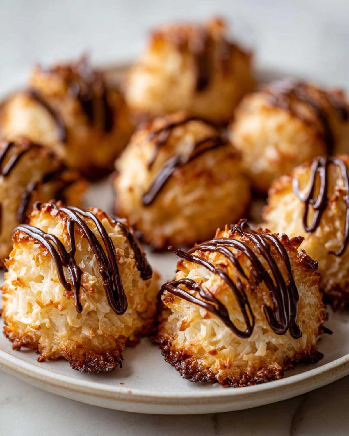 Close-up of golden-brown Coconut Macaroons with Dark Chocolate Drizzle stacked on a plate.