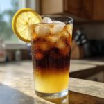 A tall glass showing layered Cold Brew Lemonade with ice, a lemon slice garnish, and condensation.