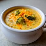 Close-up of a creamy bowl of Copycat Broccoli Cheddar Soup topped with shredded cheddar and fresh broccoli florets.
