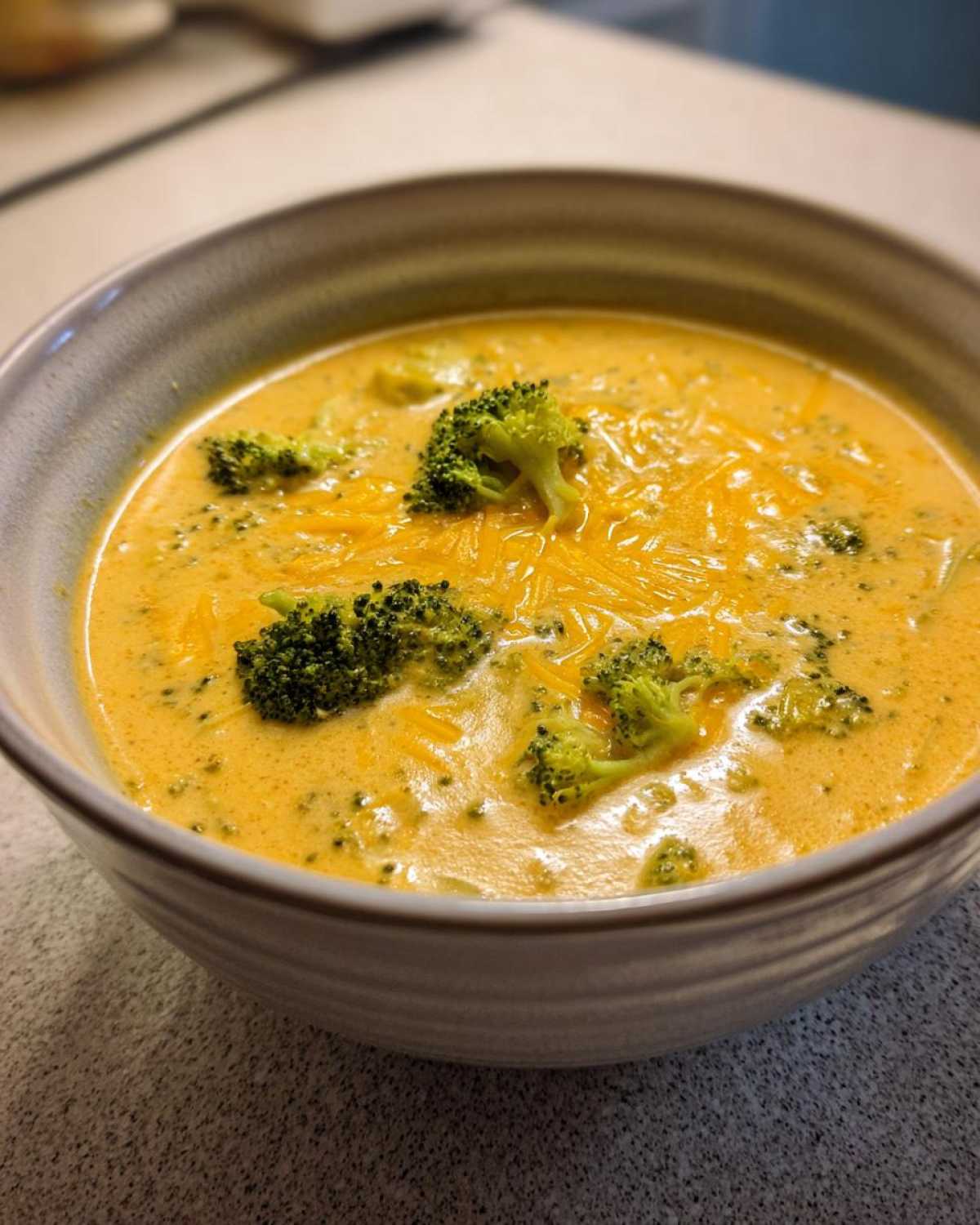 A close-up of a bowl filled with creamy Copycat Broccoli Cheddar Soup, topped with melted shredded cheddar and fresh broccoli florets.