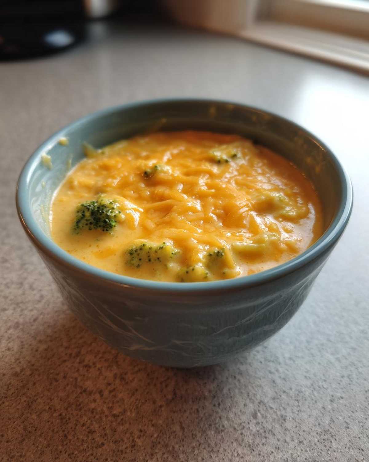 A close-up of a blue bowl filled with creamy Copycat Broccoli Cheddar Soup topped with melted shredded cheddar cheese and visible broccoli florets.