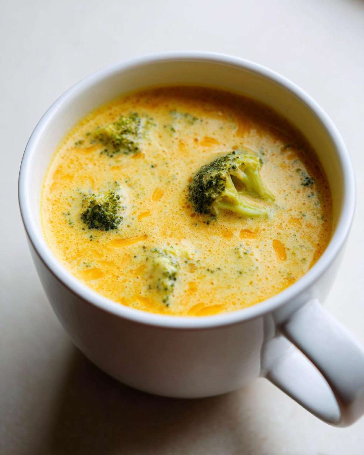 Close-up of rich, yellow Copycat Broccoli Cheddar Soup with visible broccoli florets in a white mug.