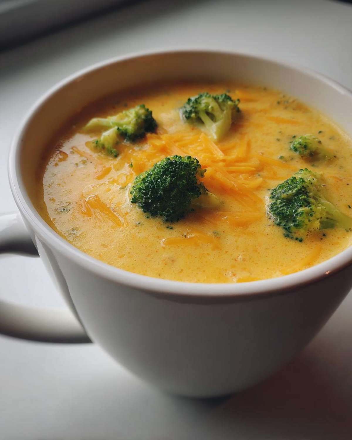 A close-up of creamy, orange Copycat Broccoli Cheddar Soup topped with fresh broccoli florets and shredded cheddar cheese.
