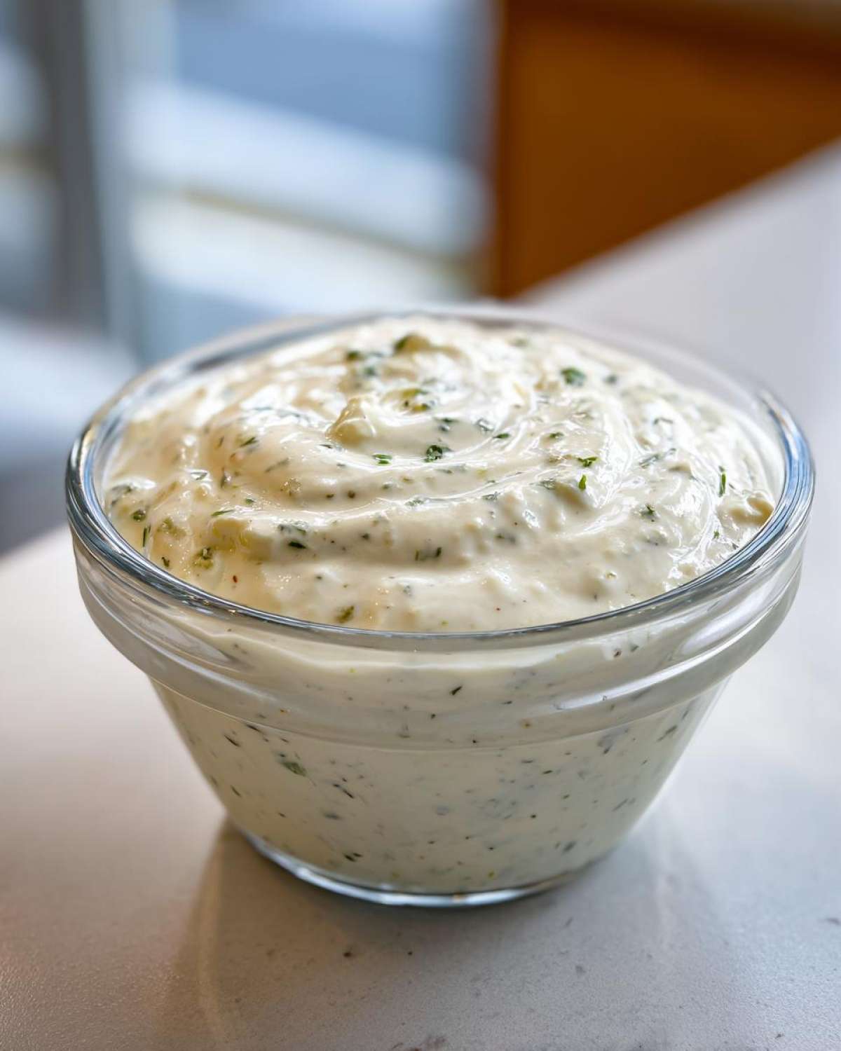 Close-up of creamy, white Copycat Olive Garden Salad Dressing speckled with green herbs in a clear glass bowl.