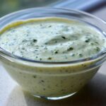 Close-up of creamy, pale green Copycat Olive Garden Salad Dressing speckled with herbs in a clear glass bowl.