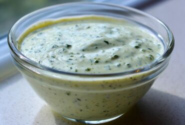Close-up of creamy, pale green Copycat Olive Garden Salad Dressing speckled with herbs in a clear glass bowl.