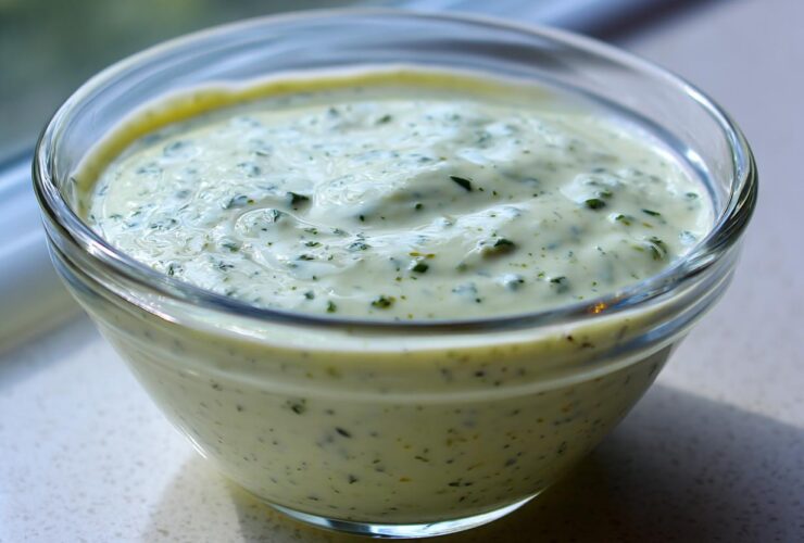 Close-up of creamy, pale green Copycat Olive Garden Salad Dressing speckled with herbs in a clear glass bowl.