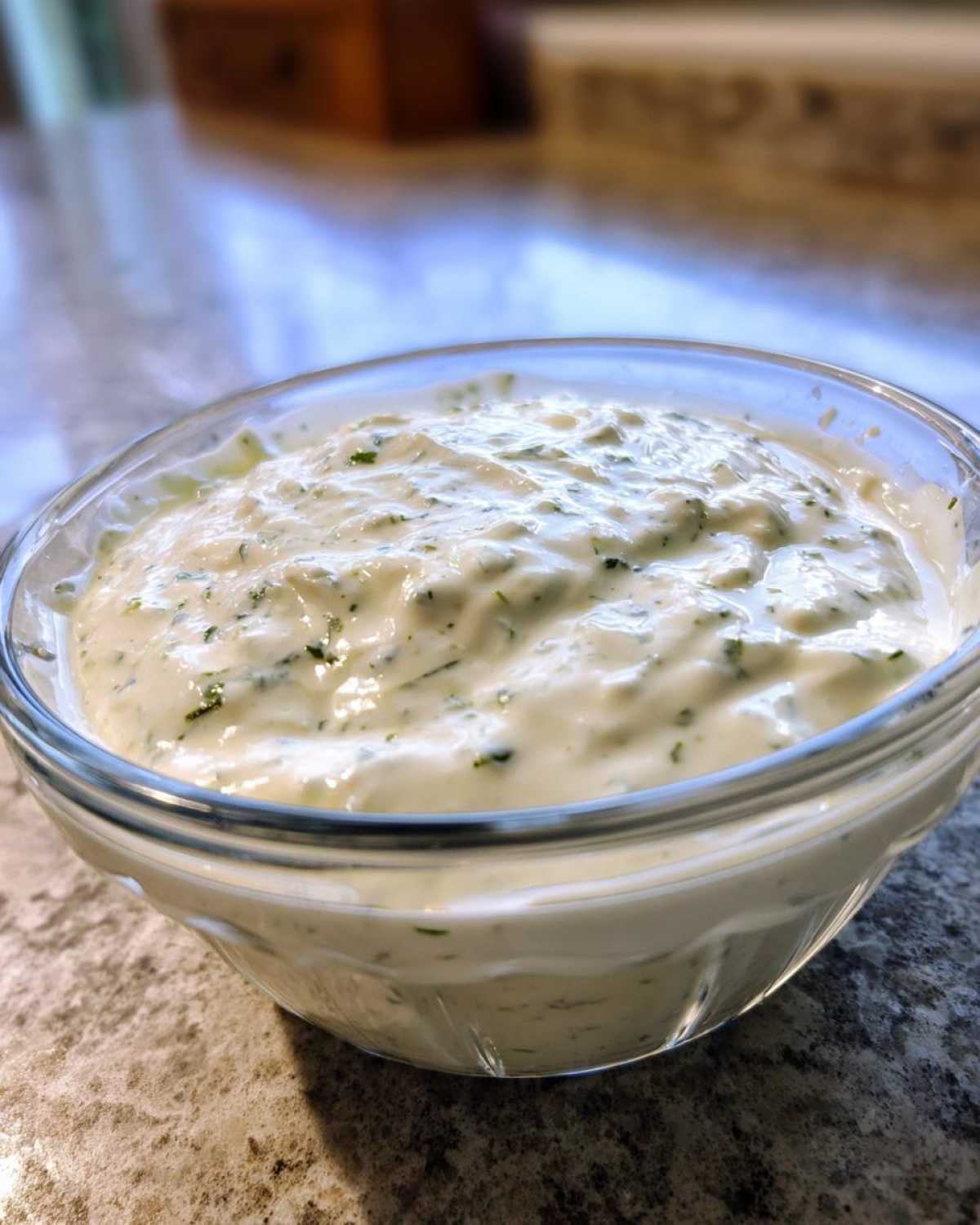 Close-up of creamy Copycat Olive Garden Salad Dressing mixed with visible green herbs in a clear glass bowl.