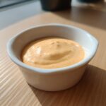 A small white ramekin filled with creamy, orange-hued Copycat Raising Cane’s Sauce sitting on a wooden table.