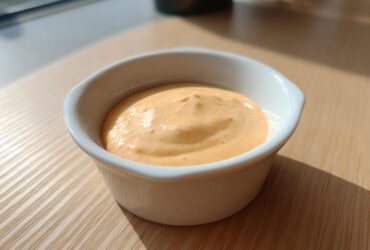 A small white ramekin filled with creamy, orange-hued Copycat Raising Cane’s Sauce sitting on a wooden table.