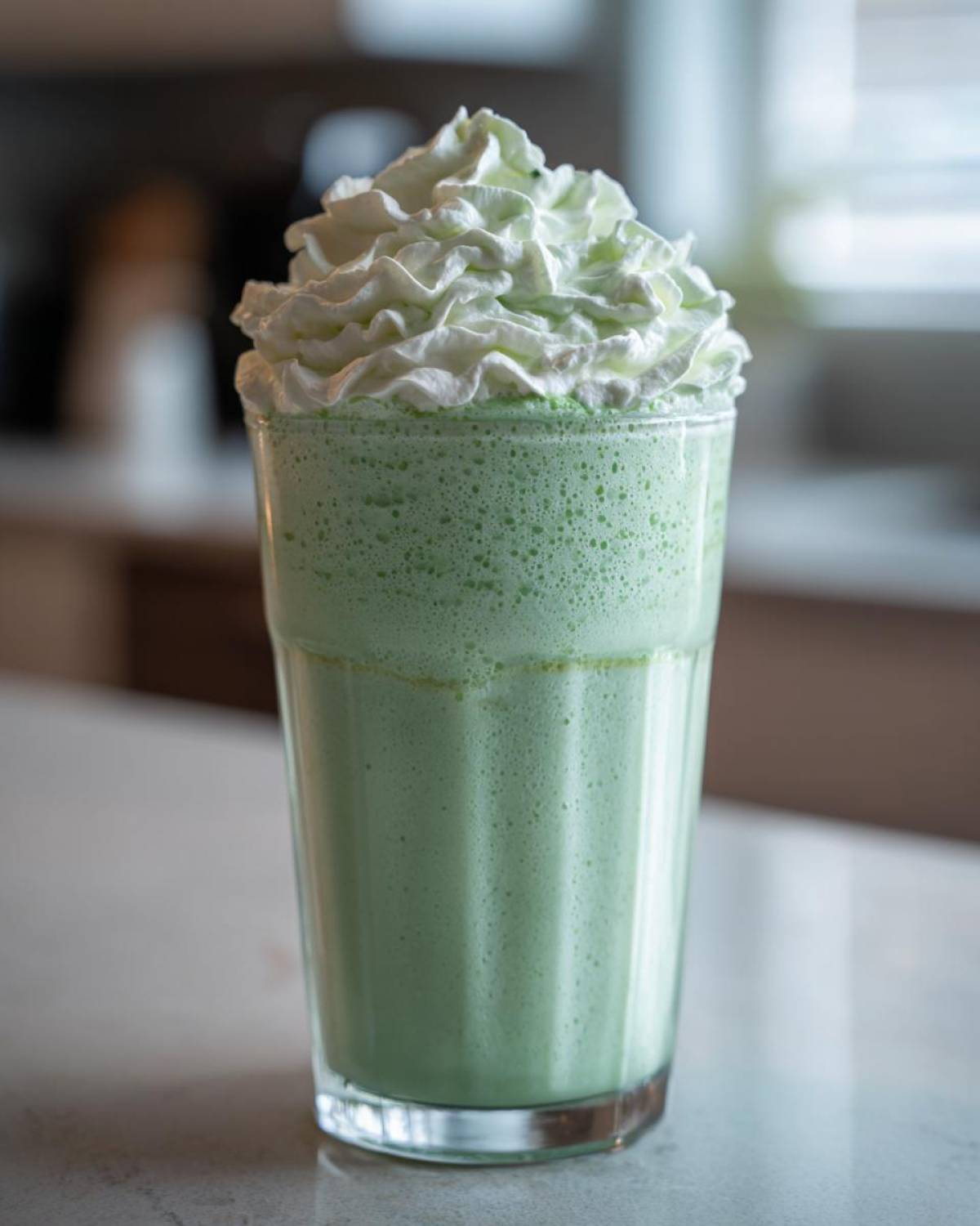 A tall glass filled with a vibrant green Copycat Shamrock Shake topped high with white mint whipped cream.