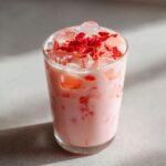 Close-up of a tall glass filled with a pink, creamy Copycat Starbucks Pink Drink, topped with ice and dried red fruit pieces.