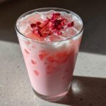 A tall glass filled with a creamy pink beverage, ice, and topped with dried rose petals, representing the Copycat Starbucks Pink Drink.