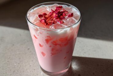 A tall glass filled with a creamy pink beverage, ice, and topped with dried rose petals, representing the Copycat Starbucks Pink Drink.