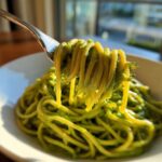 A fork lifting a generous swirl of bright green Creamy Avocado Pasta from a white bowl.