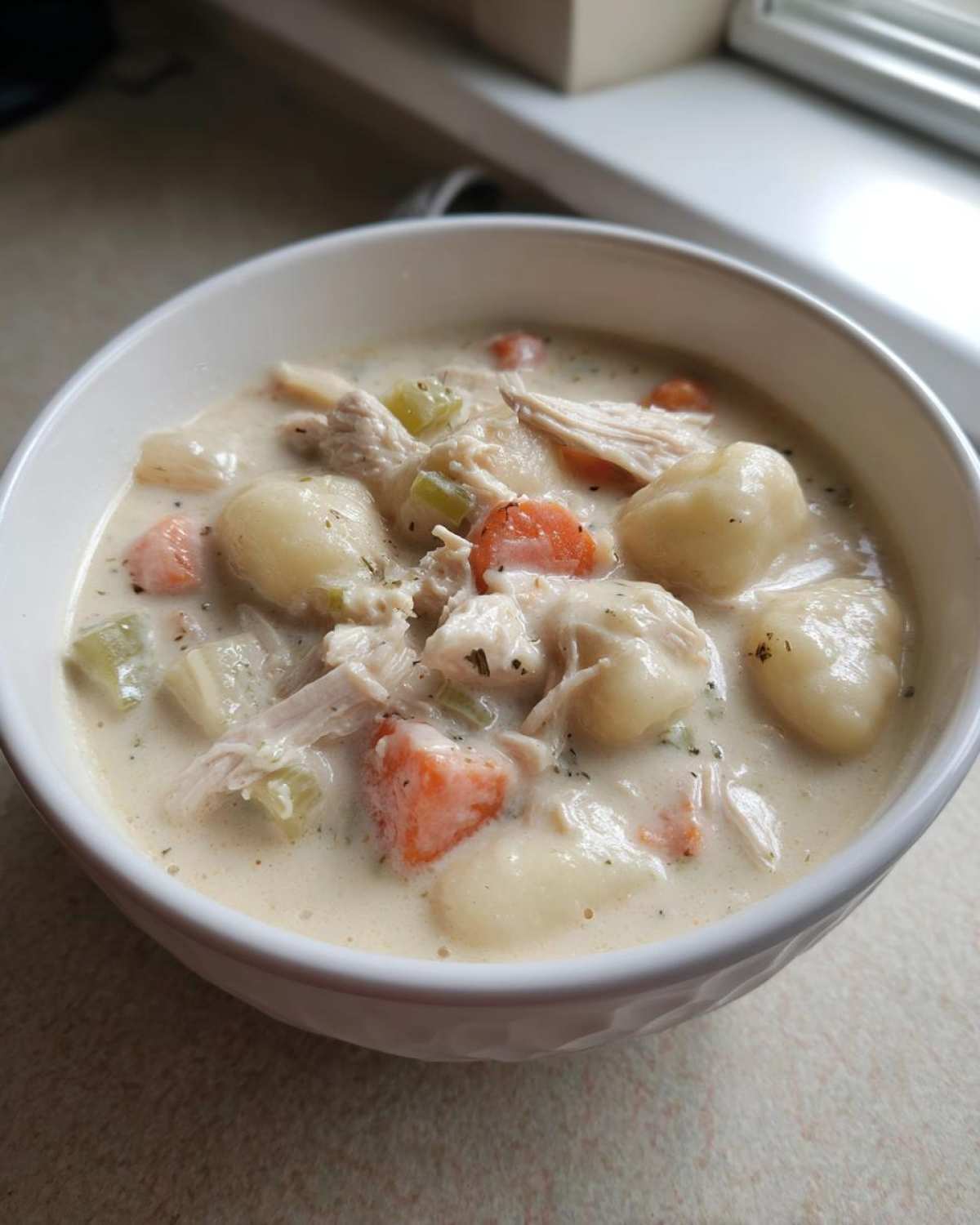 A close-up of a white bowl filled with rich Creamy Chicken Gnocchi Soup, featuring shredded chicken, carrots, celery, and soft gnocchi.