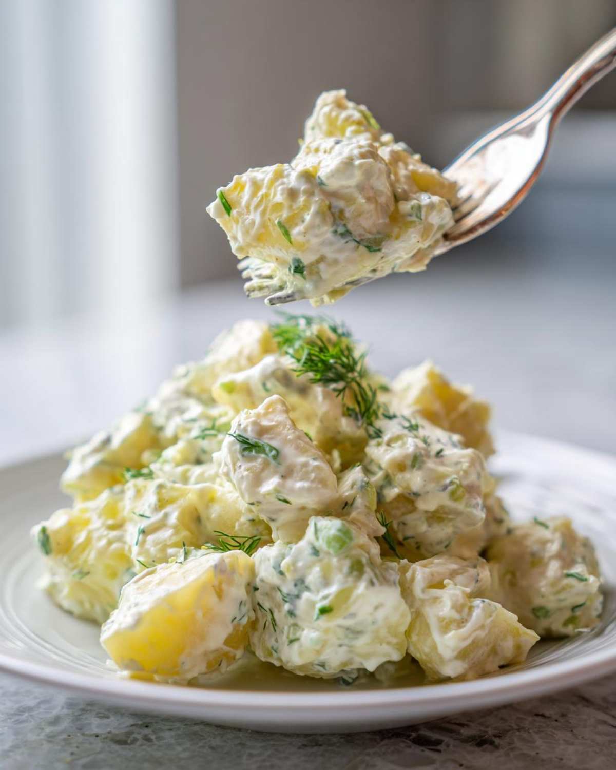 A fork lifts a bite of rich, creamy dill potato salad from a white plate piled high with the salad.