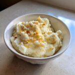 A white bowl filled with fluffy Creamy Garlic Mashed Cauliflower topped with browned garlic bits.