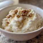 A white bowl filled with fluffy Creamy Garlic Mashed Cauliflower, topped with chopped nuts and a drizzle of liquid.