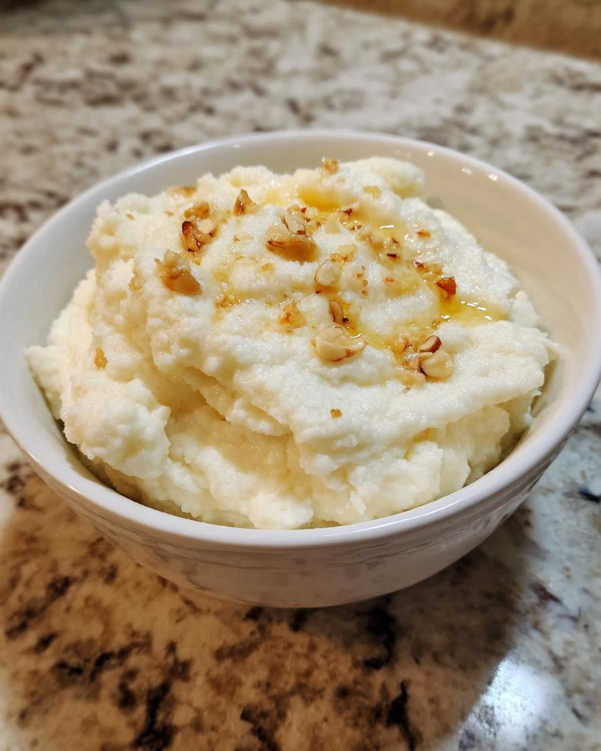A white bowl filled with fluffy Creamy Garlic Mashed Cauliflower topped with chopped nuts and a drizzle of oil or butter.