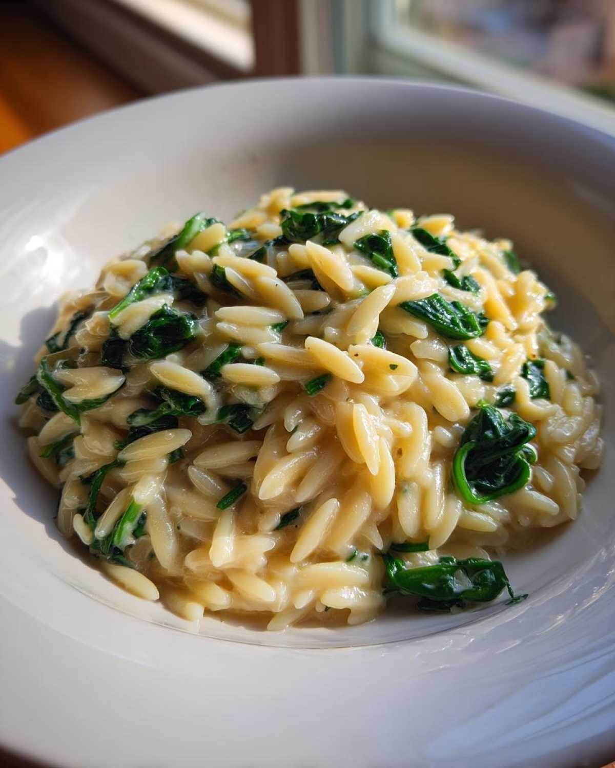 A close-up of creamy Garlic Parmesan Orzo with Spinach served in a white bowl, beautifully lit by sunlight.