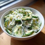 Close-up of a white bowl filled with slices of Creamy German Cucumber Salad, coated in a white dressing and topped with fresh dill.