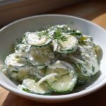 A white bowl filled with slices of Creamy German Cucumber Salad, coated in a white dressing and garnished with fresh dill.