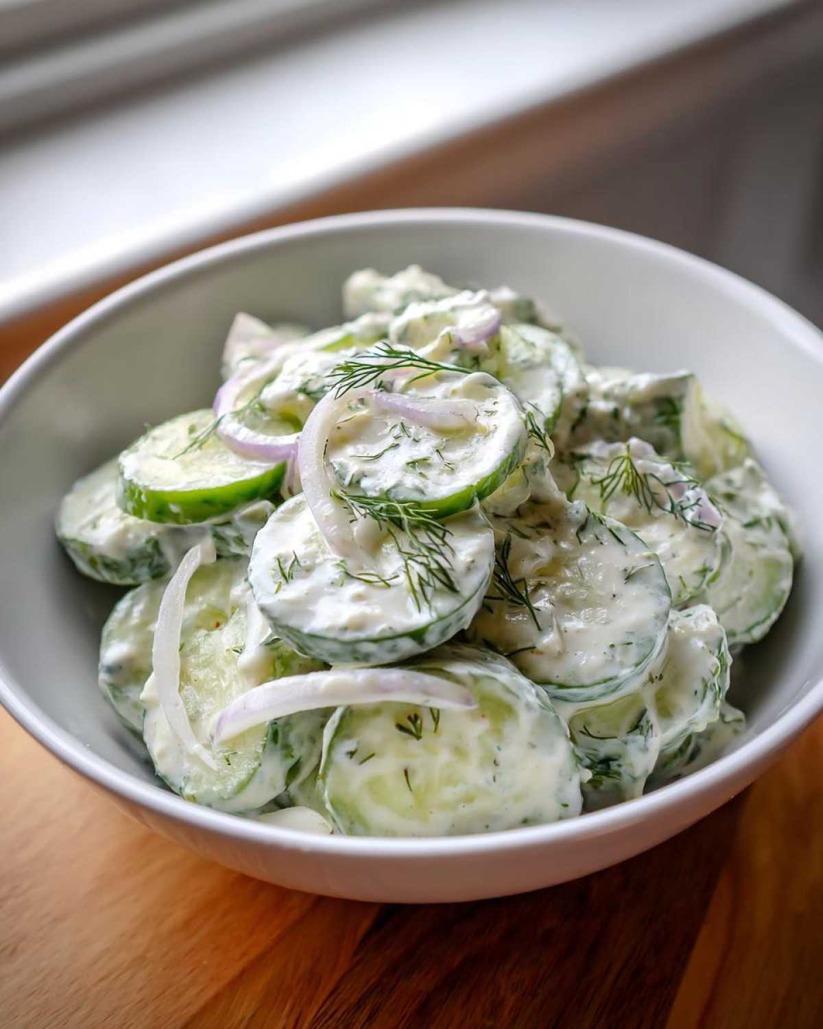 Close-up of a white bowl filled with Creamy German Cucumber Salad, featuring sliced cucumbers, creamy dressing, red onion, and fresh dill.