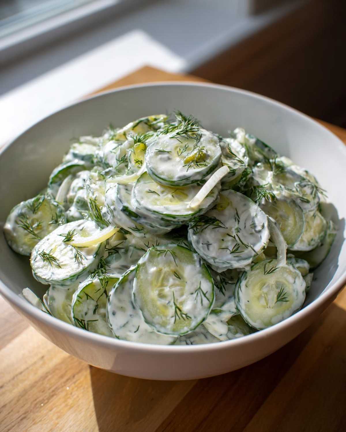 A close-up of a white bowl filled with Creamy German Cucumber Salad, featuring sliced cucumbers coated in a white dressing and topped with fresh dill.